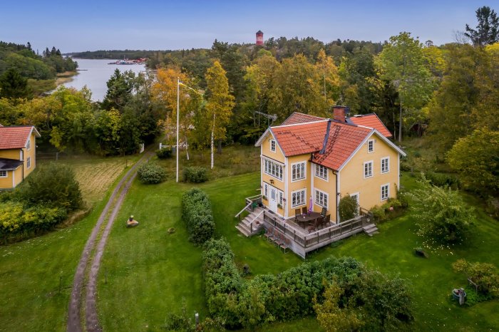 Flygbild av ett tvåvånings gult hus med rött tegeltak, omgivet av grönskande trädgård och natur.