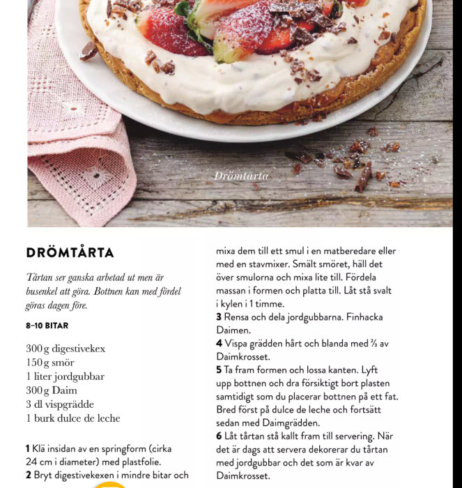 Bild av receptet på en drömtårta garnerad med jordgubbar och Daim, serverad på en tallrik med en träbakgrund.