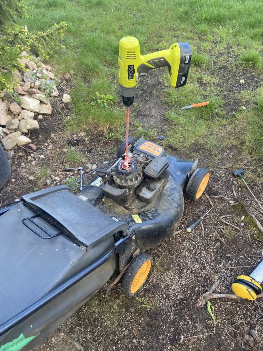 En skruvdragare används för att starta en gräsklippare med öppen motorhuv och synliga delar på en gräsmatta.