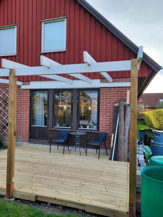 Pågående bygge av träaltan med fyra takbjälkar monterade framför ett rött hus med vita fönsterkarmer.