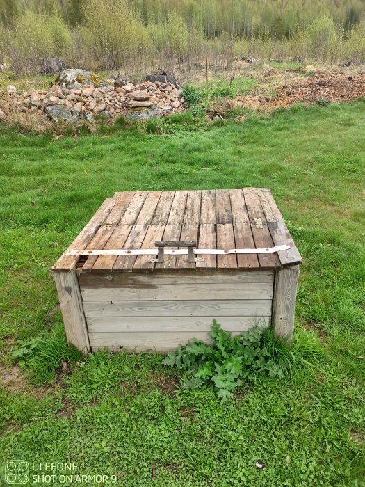 Tungt, delat lock på en kompost med gångjärn, omgiven av grönt gräs i en trädgård.