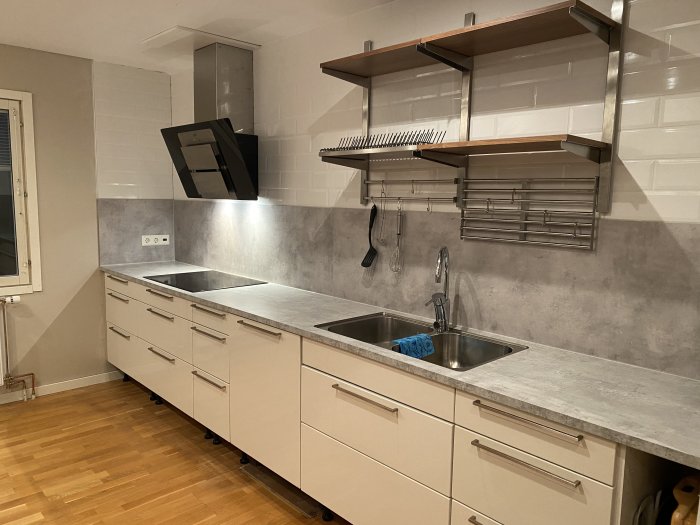 Kök med vita skåp, rostfri diskho och öppna trähyllor på en kaklad vägg, redo för belysningsinstallation.