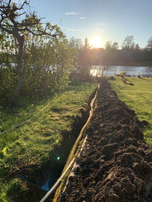 Grävd ränna i en trädgård vid solnedgång med en placerad slang som sträcker sig mot sjön i bakgrunden.