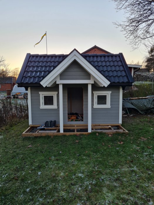 Gråmålad lekstuga under konstruktion med svart tak, två fönster och en svensk flagga på taket, placerad på en gräsmatta.