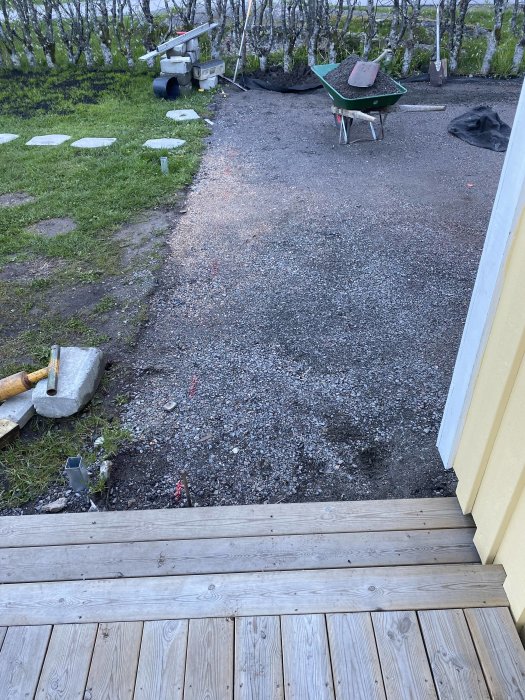 Uppfart under förberedelse för asfaltering, angränsande till träaltan, med skottkärra och byggmaterial.