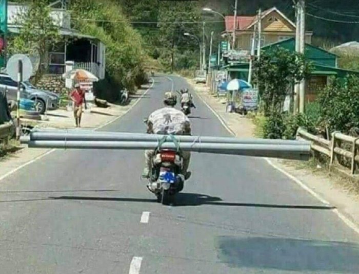 Motorcykel med långa rör fastsurrade på sidorna som sträcker sig bortom fordonets bredd, på en väg.