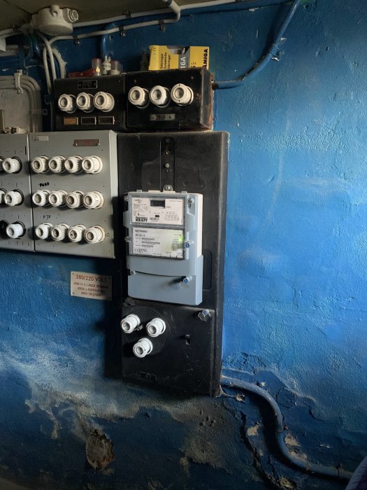 Elektrisk installation med säkringsskåp, mätare och kablar på en blå vägg.