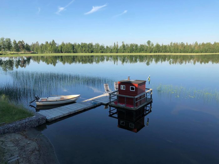 Träbrygga med båt och bastuflotte i lugn insjö omgiven av skog och klar himmel.