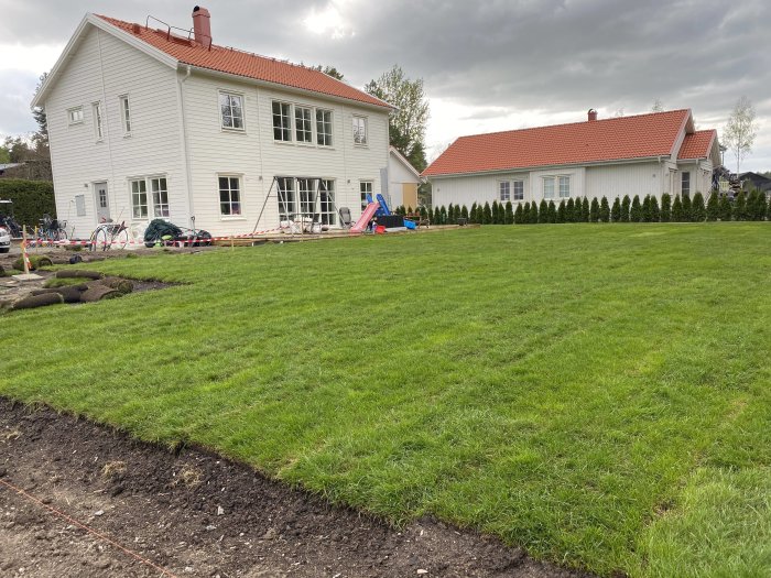 Nyplanterad gräsmatta med rullat gräs framför en vit villa, gräsmattejord och lekplats i bakgrunden.