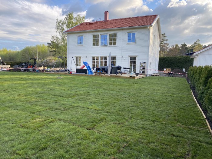 Nylagd gräsmatta med jämn grön yta framför en vit villa, utrustning synlig i bakgrunden.