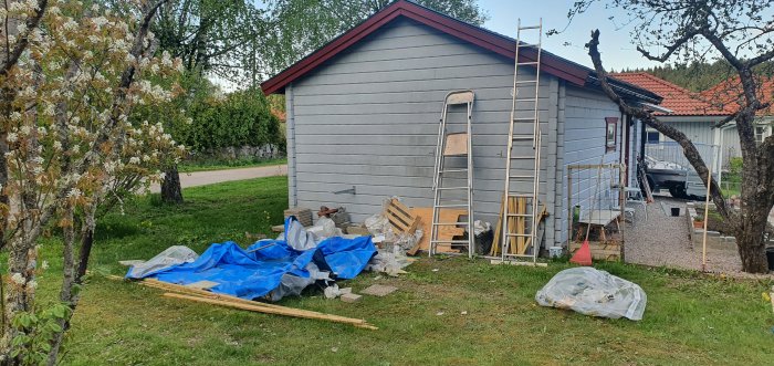 Hög med byggspill framför nyrenoverat hus, stegar mot vägg och gräs, delvis täckt av blå presenning.