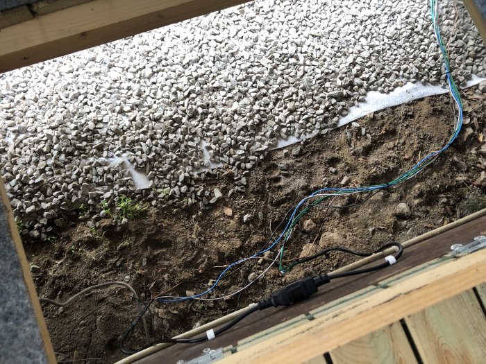Elektriska kablar och en kontakt lödda utomhus nära grus och jord under en träkonstruktion.