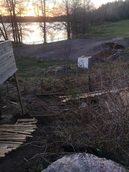 Byggarbetsplats med trallvirke på grus och sjövy i skymningen.