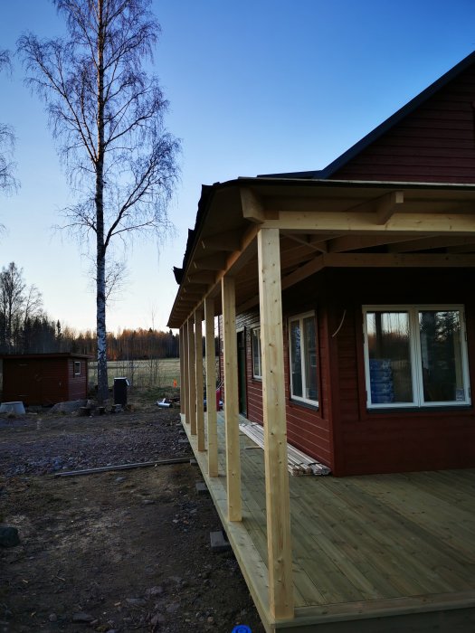 Nybyggd träveranda med takfot och hörnvinkel vid ett rödmålat hus, omgiven av natur och en björk.