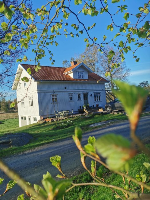 Ett tvåvånings trähus med röda takpannor sett genom grönskande trädkvistar i solljus.