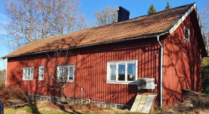 Rödmålad äldre fastighet med slitet tegeltak och synliga takstolar mot en bakgrund av lövträd och klarblå himmel.