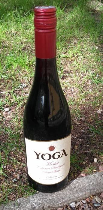 Flaska med rödvin märkt "YOGA" står på grus och gräs.