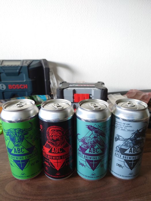 Fyra ölburkar från Apex Brewing Company med olika färg etiketter och konstverk framför en verktygslåda.