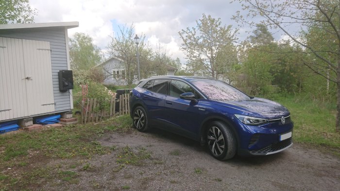 Blå elbil parkerad framför hus med laddstation, indikerar att bilen laddas.