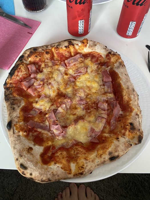 Hemlagad pizza med surdeg, skinka och smält fulost på vit tallrik, två Colaburkar i bakgrunden.