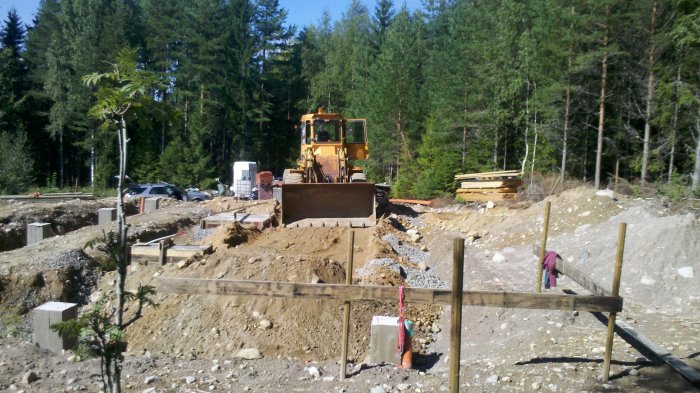 Krypgrund under konstruktion med betongplintar och balkar, grävning i skogsmiljö, hjullastare i bakgrunden.