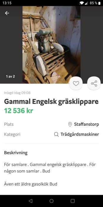 Gammal engelsk gräsklippare i ett skjul, rostig med gröntad finish, placerad bland andra föremål.