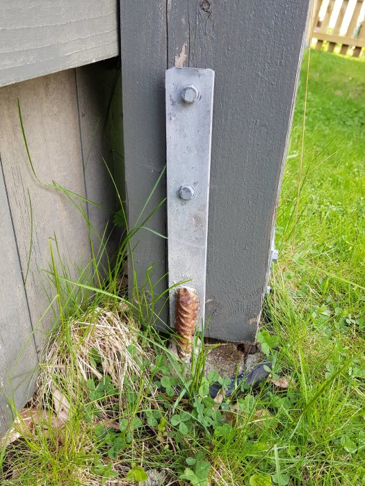 En metalforstærkning fastgjort til en træbjælke ved jorden med to bolte, omgivet af græs.