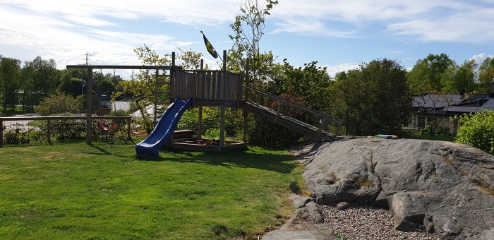 Trädäck med staket 140 cm högt över en sandlåda, rutschkana på sidan, och svensk flagga i bakgrunden, omgivet av grönt gräs.