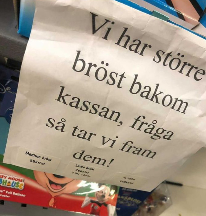 Skylt med texten "Vi har större bröst bakom kassan, fråga så tar vi fram dem!" ovanför en prislista.