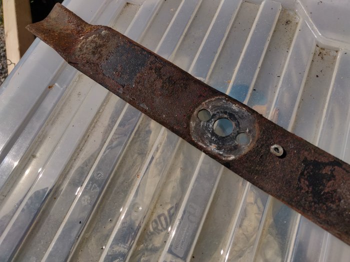 Rostig gräsklipparblad med slitet centrumhål och rester av en låssprint som ligger på en plastback.