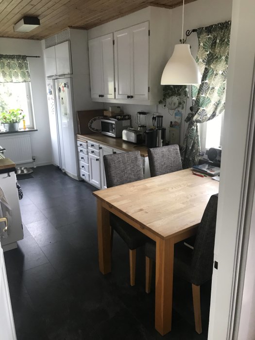 Ett mindre kök med vit skåpinredning, matplats, mörkt golv och gröna gardiner.