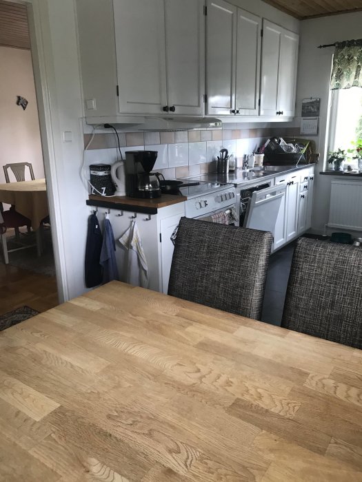 Kök med vit skåpinredning och träbänkskiva, synlig matplats och fönster.