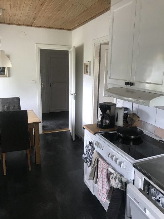 Köksinteriör med vit spis, diskmaskin, skåp och en matplats med bord och stol, träbjälkar i taket.