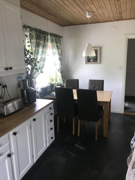 Trångt kök med vit inredning, matbord och stolar framför fönster med gröna gardiner, träbjälkar i taket.