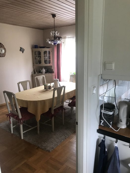 Matrum med träbord, stolar med röda kuddar, grå matta, vita skåpet och ljuskrona, sedd genom en dörröppning.