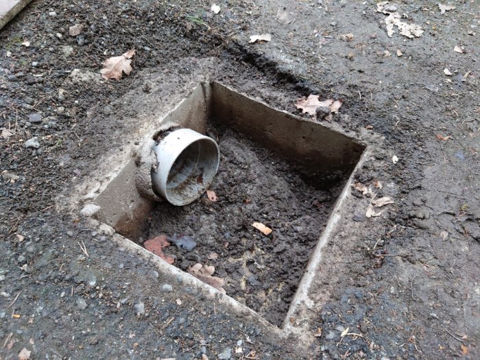 Trasigt dagvatten-galler vid garage, med synligt 110mm plaströr och betongrör i hål fyllt med sten och jord.