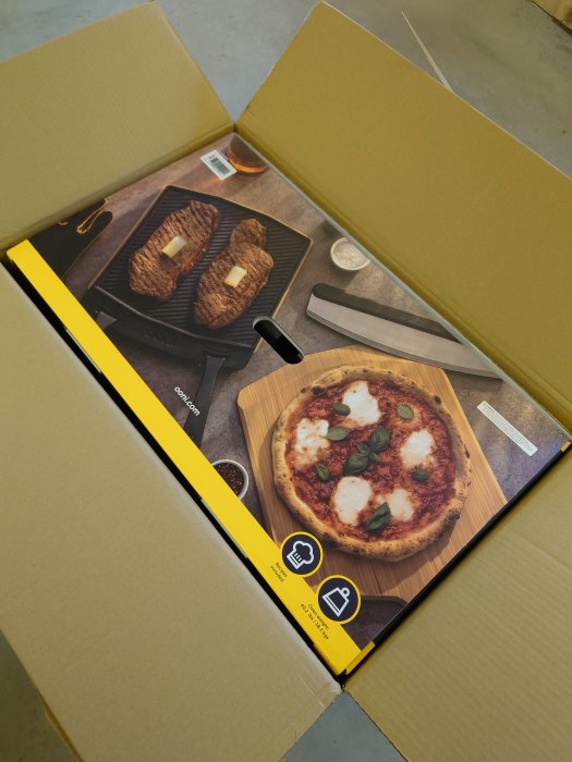 Pizzasten i öppen kartong redo för unboxing, skildrar grillade biffar och pizza på förpackningen.