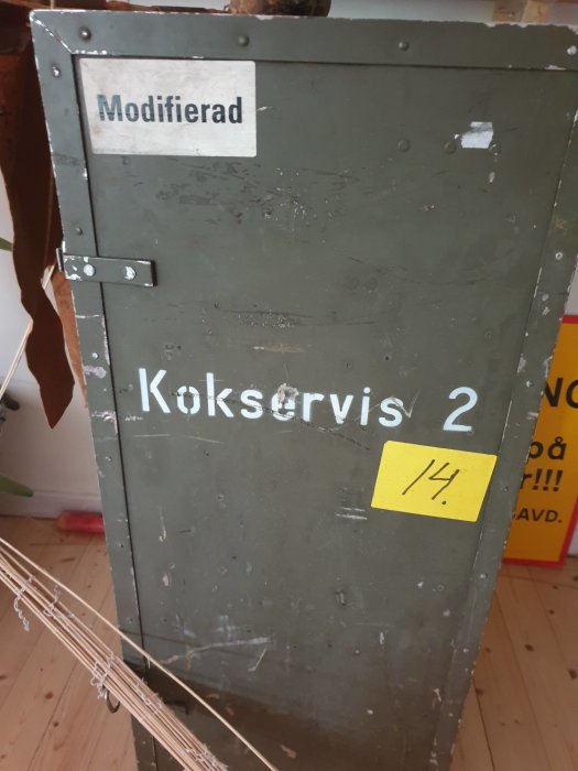 Grått metallskåp märkt "Modifierad" och "Koksservis 2", slitage och gula lappar synliga.