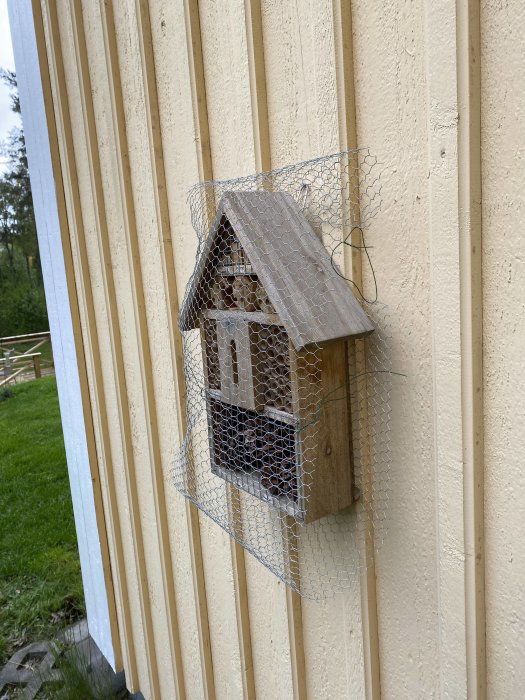 Insektshotell skyddat av hönsnät monterat på en beige husvägg för att förhindra hackspettsangrepp.