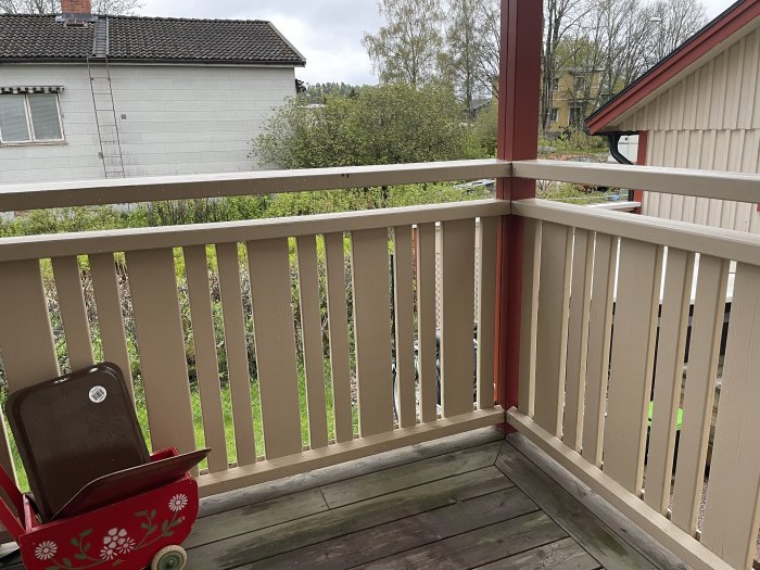 Obehandlad träveranda med räcke och vertikala spjälor framför en trädgård, med en röd lekvagn, redo för renovering.