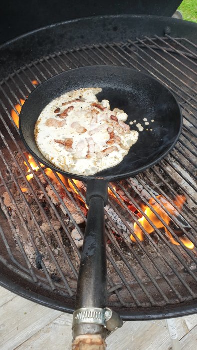 Kolbulle steks i en gjutjärnspanna över ett träkolseldat grillgaller med synliga lågor.
