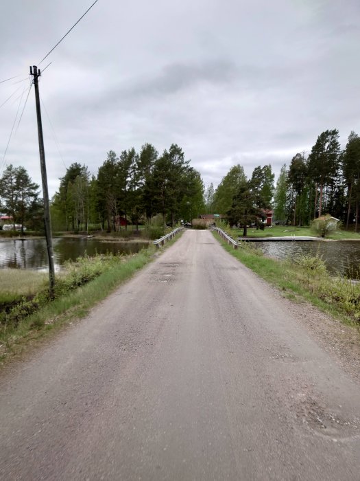 Grusväg leder över en bro med träd och elstolpar vid sidorna och en flod under broen.