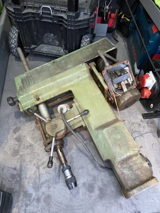 Äldre grön maskin med exponerad motor och borrchuck ligger på ett fordonsgolv bland verktygslådor.