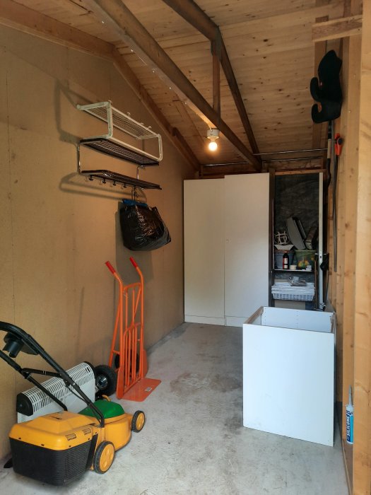 Oisolerat förråd med synliga reglar och takbjälkar, betonggolv, gräsklippare och lagringshyllor på väggen.