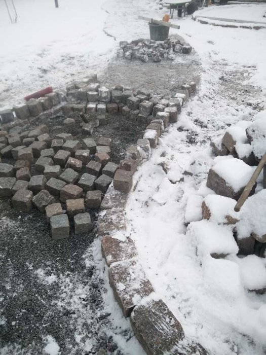 Påbörjad stensättning med smågatsten i snöväder, ofullständig med verktyg och skottkärra i bakgrunden.