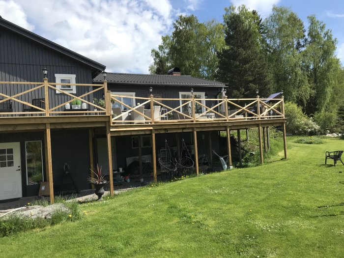 Altan av trä på mörkblått hus med staket i obehandlad trä, grön gräsmatta i förgrunden.