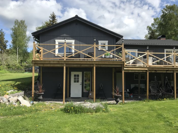 Altan på ett mörkgrått hus med trästaket och vita fönster, omgivet av grön gräsmatta.