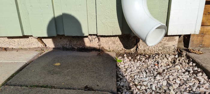Stupränna som mynnar ut nära marken ovanför en betongplatta och vita grus, tecken på vattensamling vid husvägg.