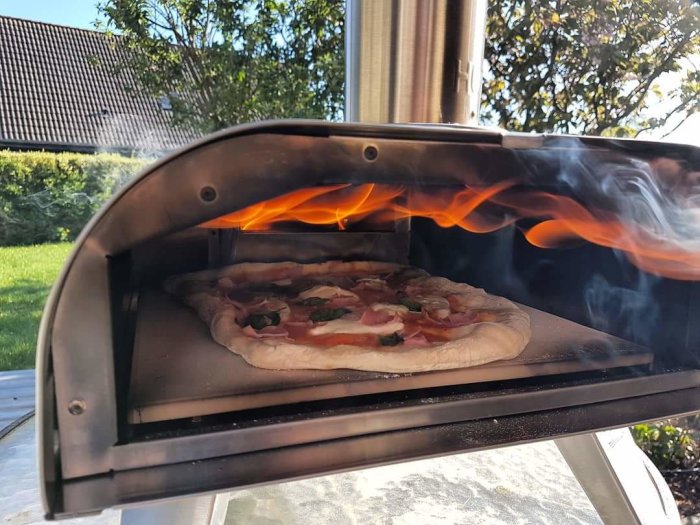 Pelletsgrill med öppen lucka där en pizza bakas, med synliga lågor och rök.