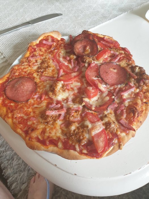 Hembakad pizza med salami, skinka och röd lök på vit tallrik, bredvid en pizzakniv.